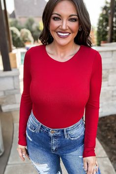 a smiling woman wearing ripped jeans and a red top is posing for the camera with her hands in her pockets