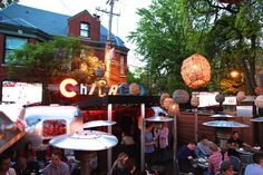 many people are sitting at tables in front of a building with lights hanging from the roof