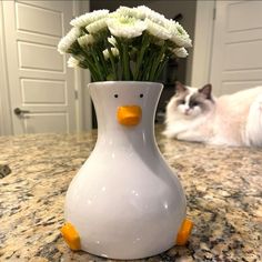 a white duck vase with flowers in it on a counter next to a gray cat