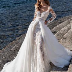 a woman in a white wedding dress standing on the rocks by the water with her arms around her waist