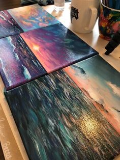 three paintings are sitting on a table next to a cup and coffee mug, one is painted with acrylic paint