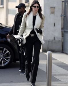 a woman walking down the street wearing black pants and white fur coat with her hands in her pockets