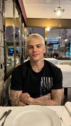 a man with tattoos sitting at a table in front of a white plate and silverware