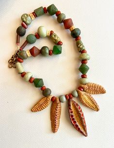 Please no discount coupons on sale items. Thank you.  The wonderfully textured stoneware leaves and pods in this necklace were hand formed and fired by another Etsy artisan.  I added color, a soft golden brown for the four smaller dangles, 35-49mm, and a deeper yellow-brown with touches of coral for the central pod, 52mm. They are strung with orange-red, green, and tan beads in a variety of shapes and materials, including old glass, ceramic, stone, and bone. Most of the beads are matte, earthy, Handmade Red Nature-inspired Necklace, Nature-inspired Jewelry With Dangling Beads For Gifts, Earthy Red Handmade Jewelry, Earthy Red Jewelry With Natural Stones, Nature-inspired Necklace With Colorful Beads For Gifts, Nature-inspired Necklace With Large Beads For Gifts, Nature-inspired Colorful Beads Necklace As Gift, Nature-inspired Colorful Beaded Necklace As Gift, Red Natural Stones Nature-inspired Jewelry