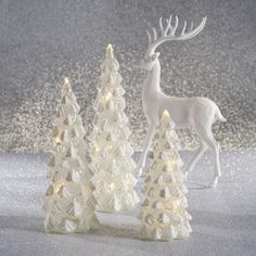 three white christmas trees with lights on them and a deer figurine in the middle