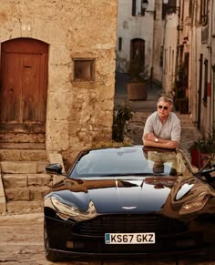 a man leaning on the hood of a black sports car in front of a cobblestone street