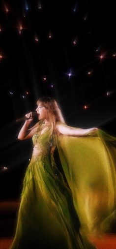 a woman in a long green dress holding a microphone and singing into a microphone with lights behind her