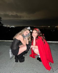 two beautiful young women dressed in costumes sitting on the ground at night with city lights behind them