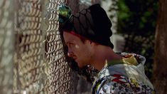 a man with a hat on leaning against a chain link fence and looking at something