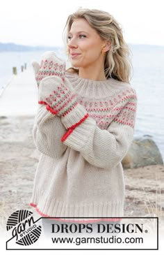 a woman standing on the beach wearing a sweater and mitts