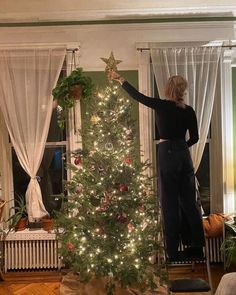 a woman decorating a christmas tree in her living room with white curtains and lights