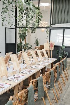 a long table with chairs and paintings on it in front of a large window filled with greenery