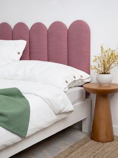 a white bed with pink headboard next to a small table and flower pot on it