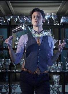 a man in a vest and tie standing next to a shelf filled with wine glasses