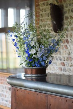 blue and white flowers are in a metal vase on a counter top next to a brick wall