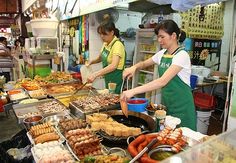 two women in green aprons cooking food at an outdoor market stall with various foods on the counter