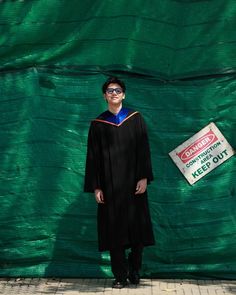 a man in a graduation gown standing next to a green tarp with a sign on it