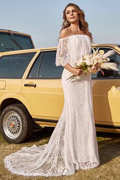 a woman standing in front of a yellow car wearing a white dress and holding flowers