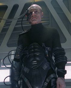 a man dressed in black standing next to another man with futuristic equipment on his chest