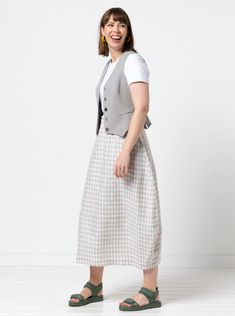 a woman standing in front of a white wall wearing a gray vest and checkered skirt