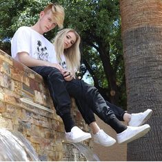 two young women sitting on top of a brick wall next to a palm tree and looking at their cell phone