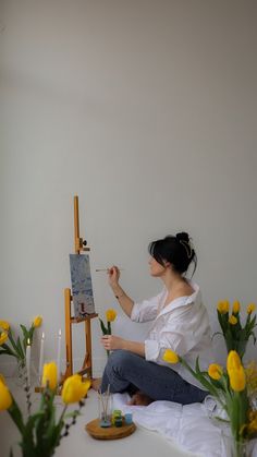 a woman sitting on top of a white table next to yellow flowers and an easel