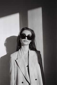 a woman wearing sunglasses and a suit standing in front of a wall with the sun shining on her face