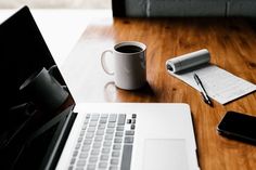 an open laptop computer sitting on top of a wooden desk next to a cup of coffee