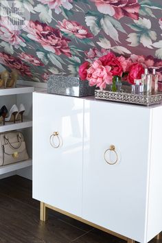 a white cabinet sitting next to a wall with pink flowers on it and two pairs of shoes