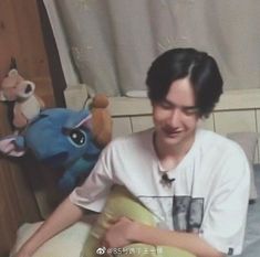 a young man sitting on top of a bed next to a stuffed animal and pillow