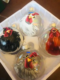 four glass christmas ornaments in the shape of roosters on a white tray with red and blue trim