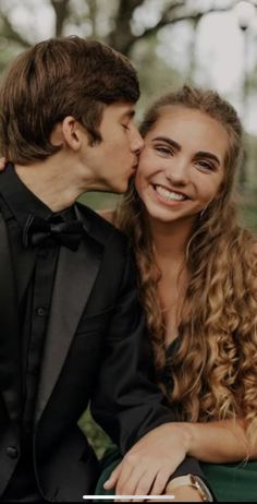 a young man and woman sitting next to each other in front of trees, kissing