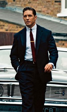 a man in a suit and tie walking next to a car with his hands in his pockets