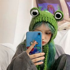 a girl with green hair wearing a frog hat and holding a cell phone up to her face