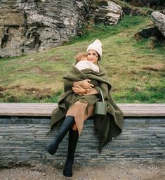 a woman sitting on top of a wooden bench next to a stone wall covered in a blanket