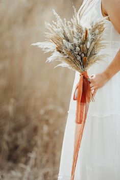 a woman in a white dress holding a bouquet of dried grass and an orange ribbon