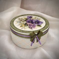 a white box with purple flowers on it and a green ribbon around the lid is sitting on a white cloth