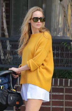 a woman in yellow sweater and white shorts walking down the street with a handbag