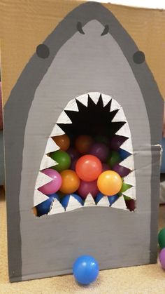 a cardboard shark with balls in it's mouth on the floor next to other toys