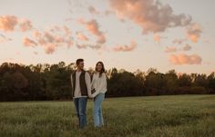 two people standing in the middle of a field with trees and clouds behind them at sunset