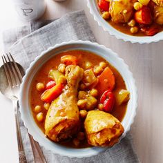 two bowls filled with stew and vegetables on top of a wooden table next to silverware