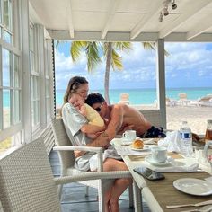 a man and woman sitting at a table in front of the ocean with breakfast on it