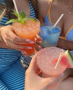 two women holding up drinks with straws and watermelon on the table in front of them