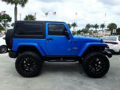 a blue jeep is parked in a parking lot with other cars and palm trees behind it