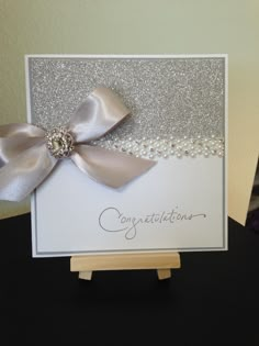 a congratulations card with a silver bow and pearls on the front, sitting on top of a wooden stand