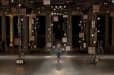 a woman walking down a runway with lots of lights on the ceiling and columns in the background