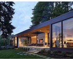 a modern house with large windows and grass in the front yard at dusk, surrounded by trees