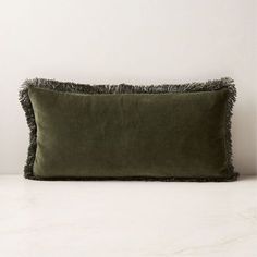 an olive green velvet pillow with fringe trim on the front and back, sitting against a white wall