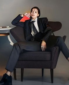 a woman in a suit sitting on a chair with her legs crossed and holding a red card