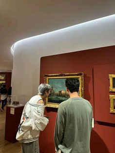 two people looking at paintings on display in a room with red walls and wooden floors